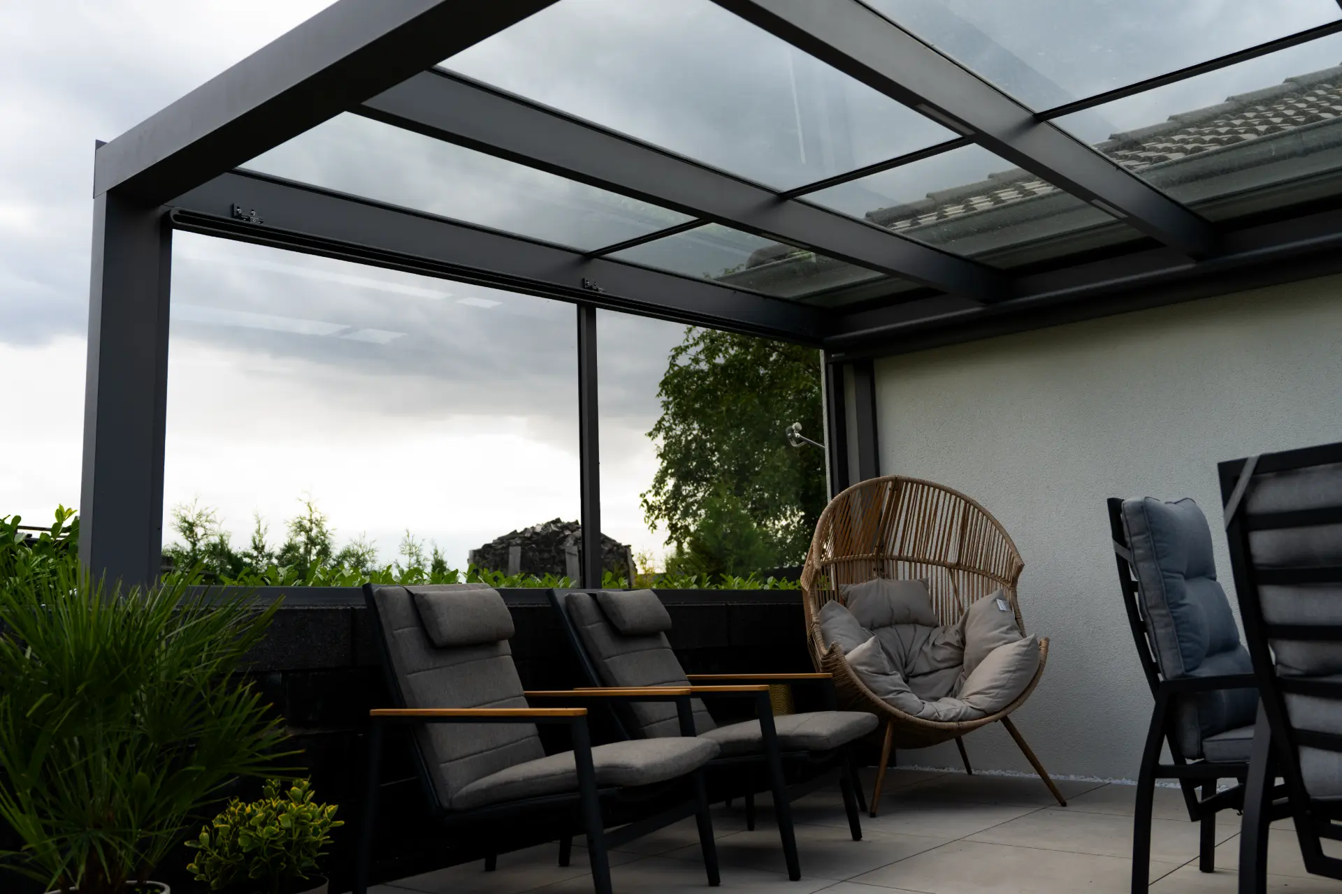 Blick von unten auf ein Terrassendach mit Glasfeldern zwischen dunklen Aluminiumsparren, seitlich eine Glasschiebewand und darunter eine Sitzgruppe