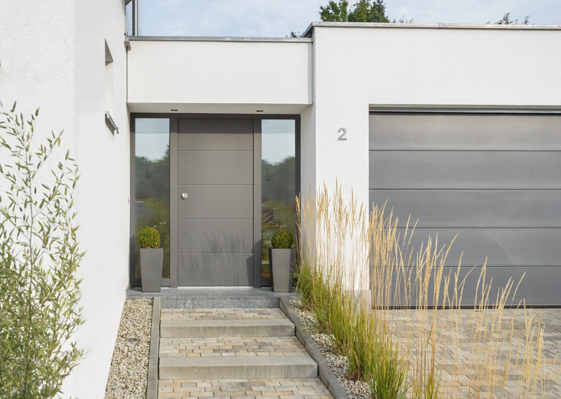 Graue Haustür mit zwei Glas-Seitenteilen neben dem passenden Garagentor, davor eine Treppe aus Pflastersteinen