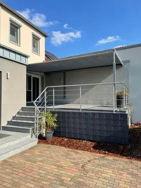 Graue Pergolamarkise auf Stützen über einer erhöhten Terrasse mit Treppe und Geländer an einem Einfamilienhaus
