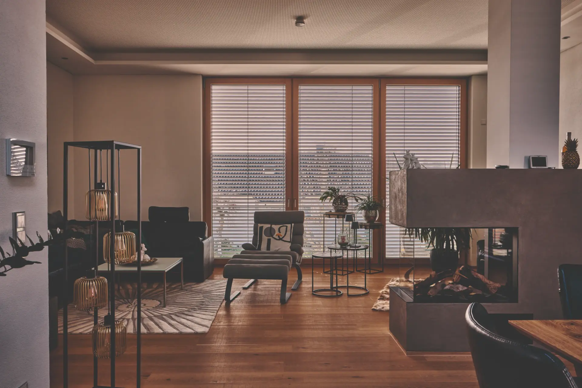 Wohnzimmer mit geschlossenen Raffstores an den Holzfenstern, davor Sessel und ein Kamin am Nachmittag