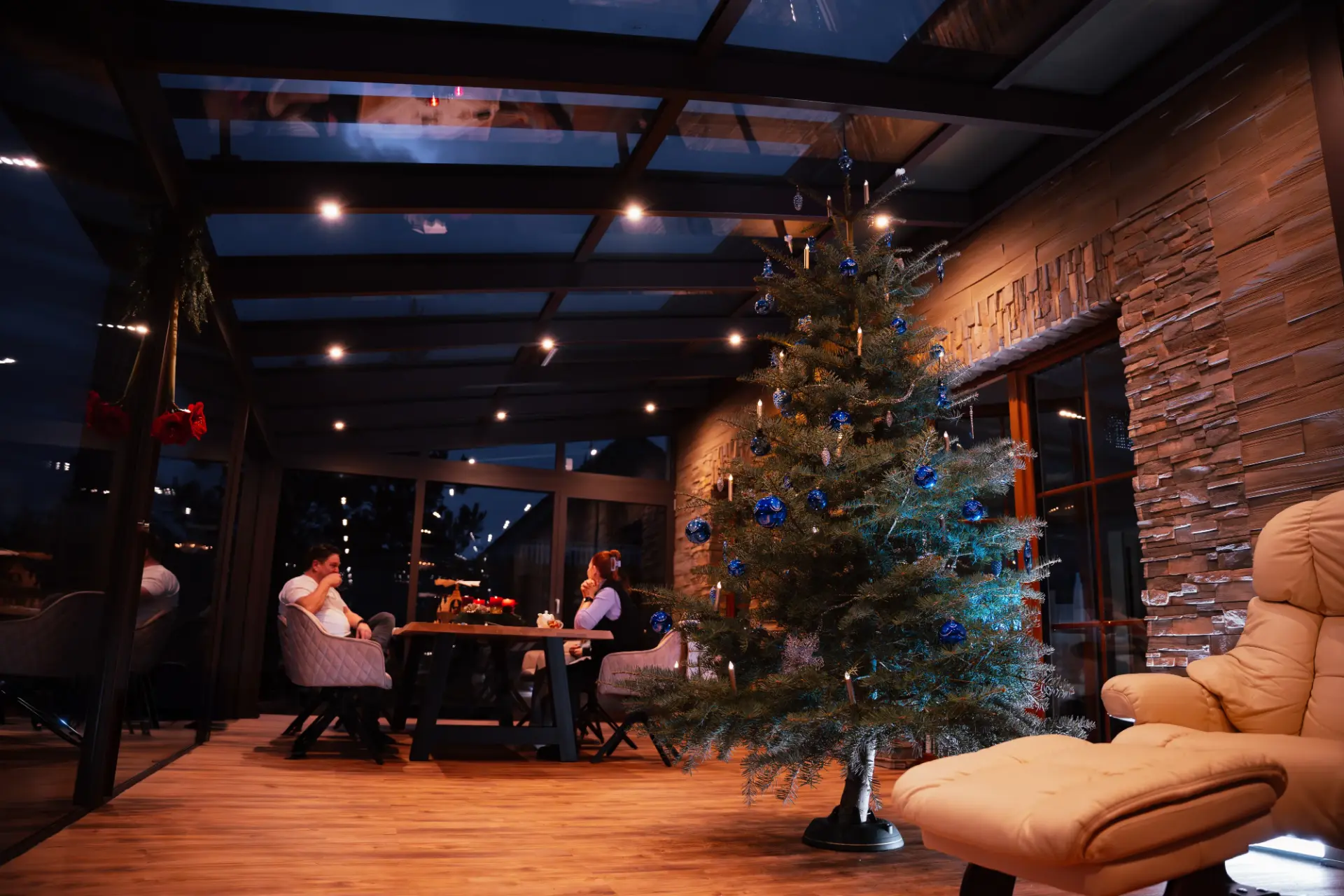 Beheizter Wintergarten am Abend mit geschmücktem Weihnachtsbaum, Esstisch und Natursteinwand