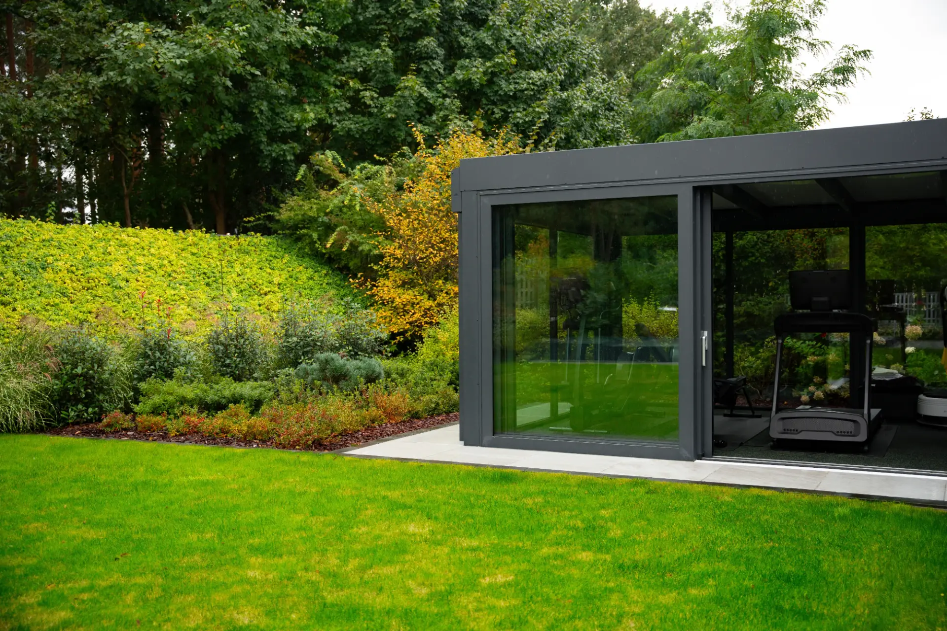 Wintergarten mit anthrazitfarbenen Aluminiumprofilen und weit geöffneter Glasschiebetür, davor Rasen und ein Staudenbeet